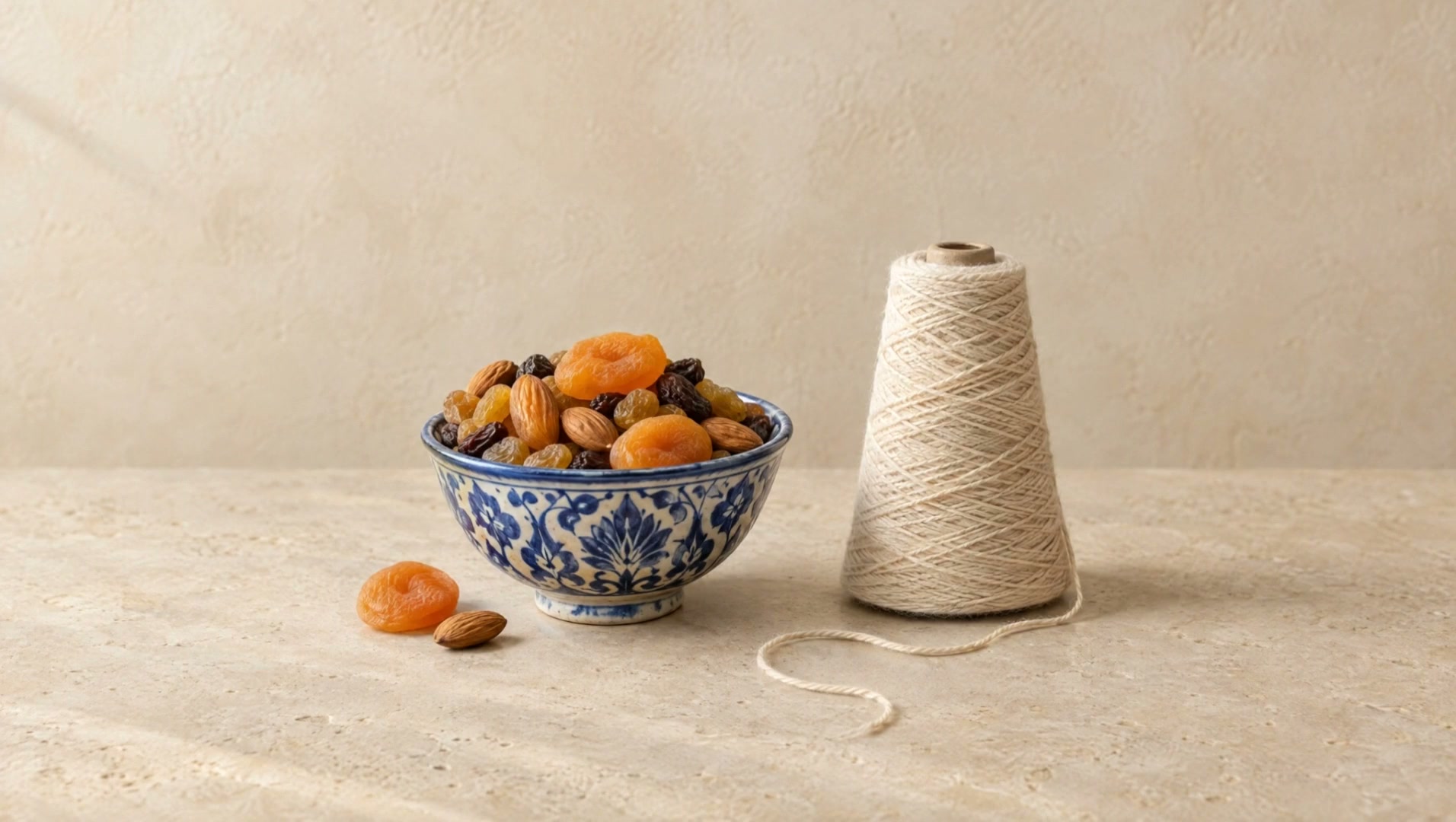A blue Rishtan bowl of dried apricots, raisins and almonds beside a cone of cotton yarn